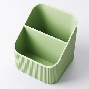 May include: A light green desk organizer with two compartments. The organizer has a rounded rectangular shape with vertical ridged detailing on the sides. The interior is smooth, and the overall design is minimalist and modern.