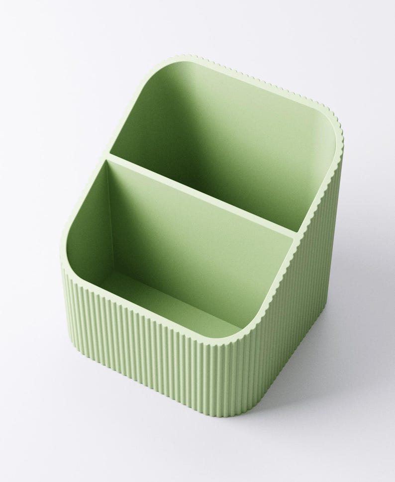 May include: A light green desk organizer with two compartments. The organizer has a rounded rectangular shape with vertical ridged detailing on the sides. The interior is smooth, and the overall design is minimalist and modern.