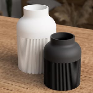 May include: Two modern vases. One is white, the other is black. Both have a cylindrical base with vertical ribbed detailing. The upper portion of each vase is smooth and rounded. The vases are displayed on a wooden surface.