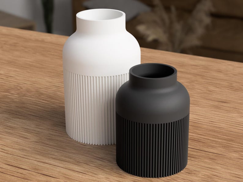 May include: Two modern vases. One is white, the other is black. Both have a cylindrical base with vertical ribbed detailing. The upper portion of each vase is smooth and rounded. The vases are displayed on a wooden surface.