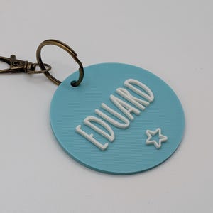 May include: A light blue round keychain with the name "EDUARD" in white, stylized letters. A small white star is next to the name. The keychain has a bronze-colored clasp.