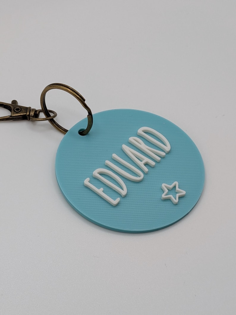 May include: A light blue round keychain with the name "EDUARD" in white, stylized letters. A small white star is next to the name. The keychain has a bronze-colored clasp.