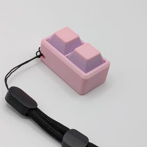 May include: A pink and purple keycap set with a black wrist strap. The keycaps are two-toned, with pink tops and purple bases. The wrist strap is black and braided, with black plastic clasps.