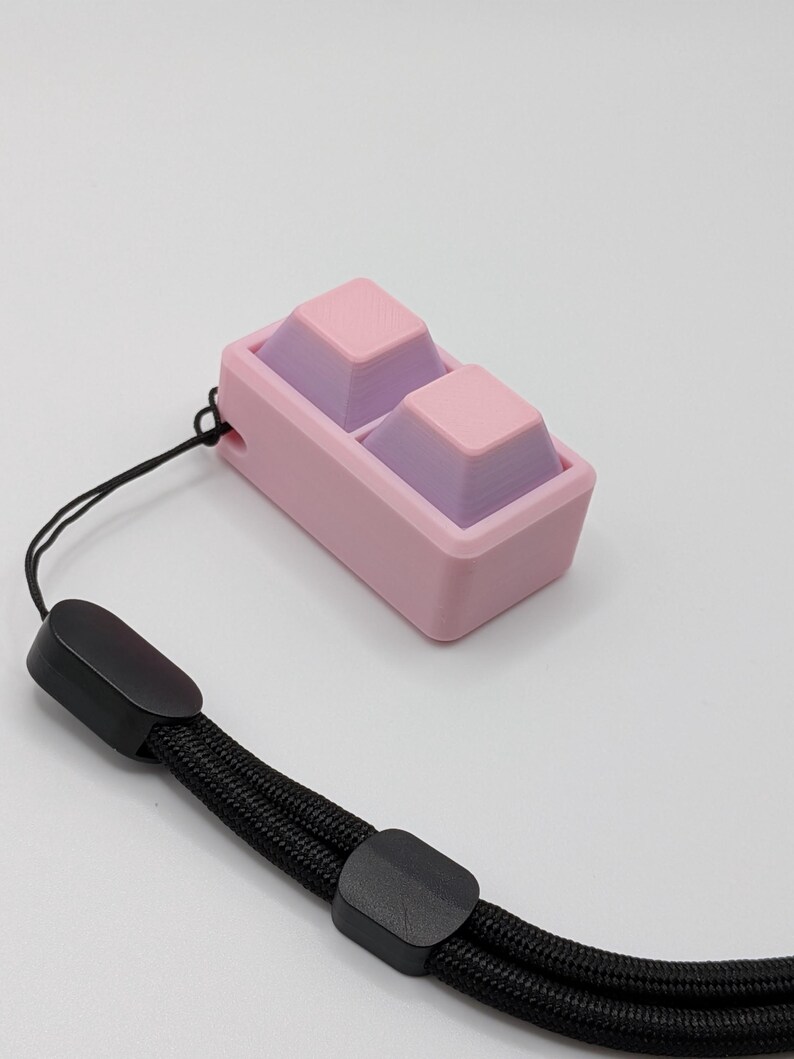 May include: A pink and purple keycap set with a black wrist strap. The keycaps are two-toned, with pink tops and purple bases. The wrist strap is black and braided, with black plastic clasps.