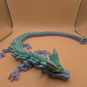 May include: A 3D-printed articulated dragon toy in shades of blue, green, and pink. The dragon has a spiky spine and crystal-like features on its head. The toy is designed to be flexible and poseable.
