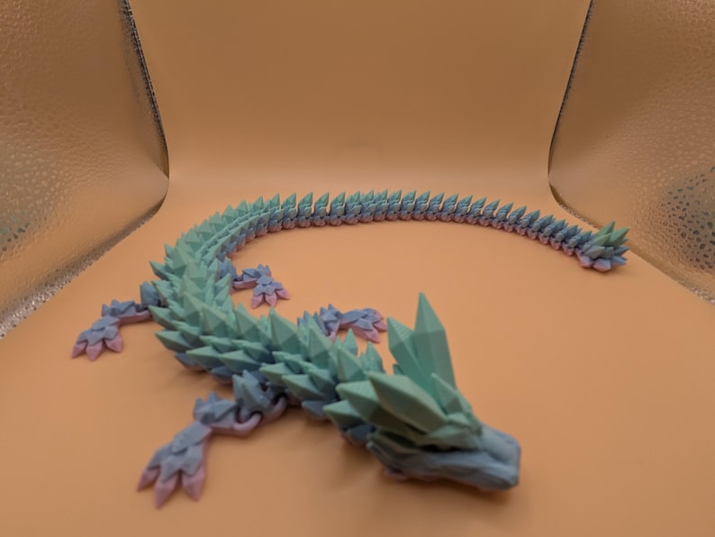 May include: A 3D-printed articulated dragon toy in shades of blue, green, and pink. The dragon has a spiky spine and crystal-like features on its head. The toy is designed to be flexible and poseable.