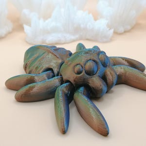 Op de afbeelding: Een 3D-geprinte spinnenfiguur met een metallic afwerking in tinten blauw, groen en bruin. De spin heeft een gedetailleerd, getextureerd oppervlak en ligt op een lichtgekleurd oppervlak. Het beeldje is een decoratief item.
