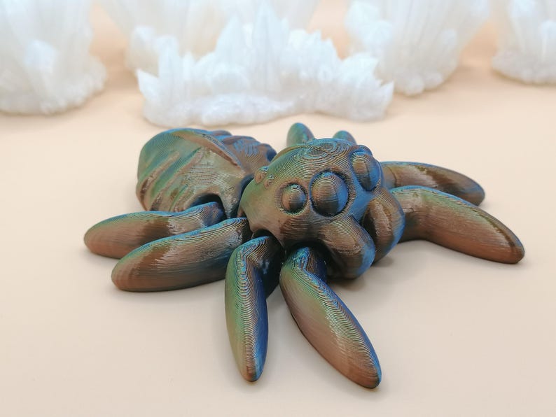 Op de afbeelding: Een 3D-geprinte spinnenfiguur met een metallic afwerking in tinten blauw, groen en bruin. De spin heeft een gedetailleerd, getextureerd oppervlak en ligt op een lichtgekleurd oppervlak. Het beeldje is een decoratief item.
