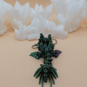 May include: A keychain featuring a detailed, iridescent green and purple coral-like design. The keychain is on a light tan surface, with several white, crystal-shaped objects in the background. The crystals have a rough, textured appearance.