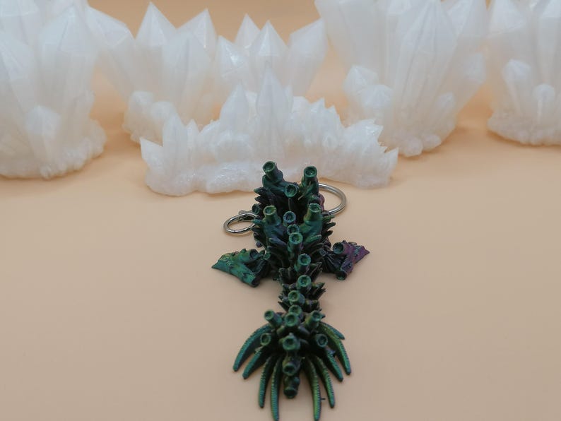 May include: A keychain featuring a detailed, iridescent green and purple coral-like design. The keychain is on a light tan surface, with several white, crystal-shaped objects in the background. The crystals have a rough, textured appearance.
