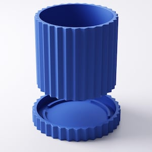 May include: A two-piece, cobalt blue container with a cylindrical body and a matching base. The body features vertical, ridged detailing, and the base has a circular design with a similar ridged edge. The container is designed to be separated from the base.