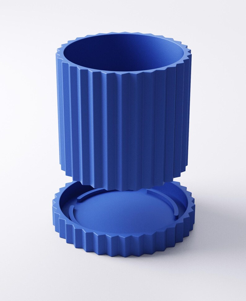 May include: A two-piece, cobalt blue container with a cylindrical body and a matching base. The body features vertical, ridged detailing, and the base has a circular design with a similar ridged edge. The container is designed to be separated from the base.