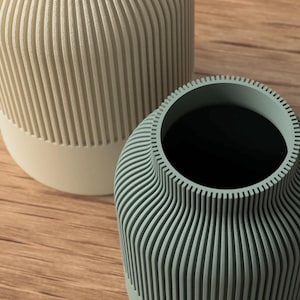 Puede incluir: Dos jarrones decorativos con patrones de estr&iacute;as verticales. Uno es de color beige claro con base blanca, el otro es de color verde azulado apagado. Los jarrones est&aacute;n sobre una superficie de madera.