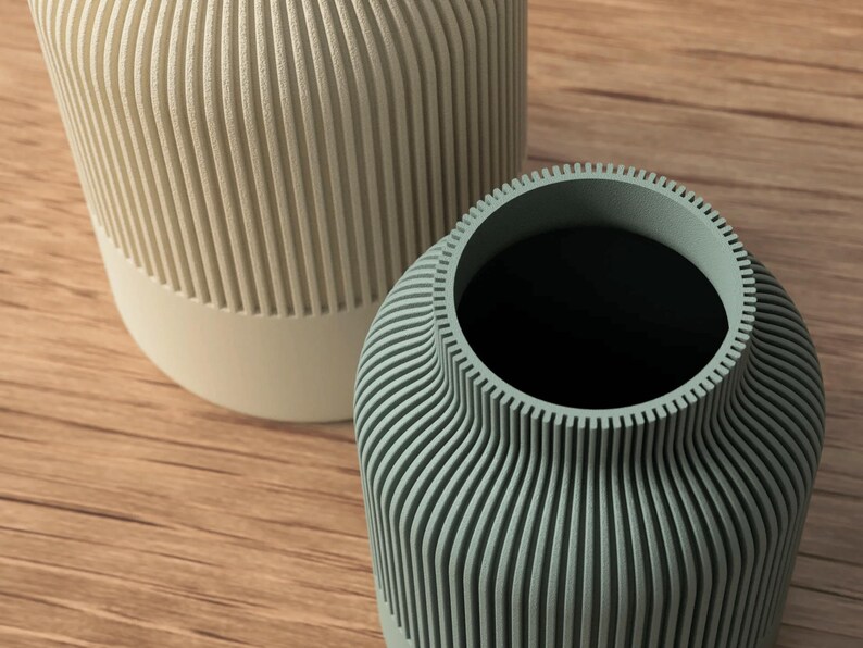 Puede incluir: Dos jarrones decorativos con patrones de estr&iacute;as verticales. Uno es de color beige claro con base blanca, el otro es de color verde azulado apagado. Los jarrones est&aacute;n sobre una superficie de madera.