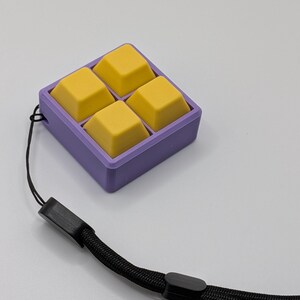 May include: A square, purple container holds four yellow, square-shaped objects. A black lanyard is attached to the container. The objects appear to be keycaps or similar components.