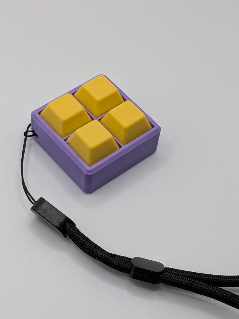 May include: A square, purple container holds four yellow, square-shaped objects. A black lanyard is attached to the container. The objects appear to be keycaps or similar components.