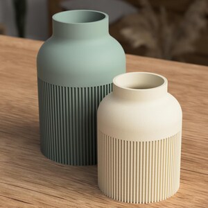 May include: Two ceramic vases, one sage green and one cream, sit on a wooden surface. The vases have a cylindrical design with vertical ribbed detailing on the lower half. The sage green vase is taller than the cream vase.