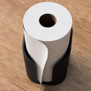 Op de afbeelding: Een zwarte papierrolhouder met een rol wit papier. Het papier is gedeeltelijk afgerold, waardoor de binnenste kartonnen kern zichtbaar is. De houder is gemaakt van donker materiaal en staat op een houten oppervlak.