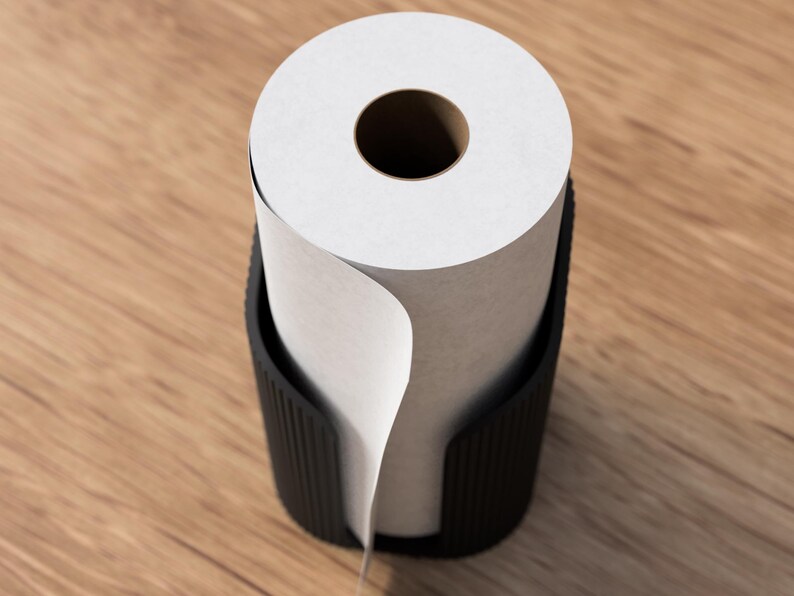 Op de afbeelding: Een zwarte papierrolhouder met een rol wit papier. Het papier is gedeeltelijk afgerold, waardoor de binnenste kartonnen kern zichtbaar is. De houder is gemaakt van donker materiaal en staat op een houten oppervlak.