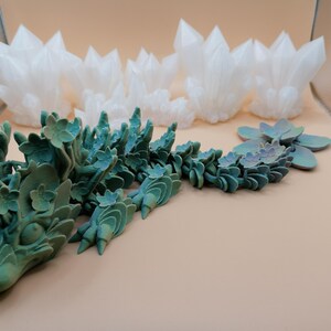 May include: A teal and green dragon sculpture with floral details and a gradient finish. The dragon is next to a cluster of white crystal formations and a succulent-like plant. The objects are on a light tan surface.