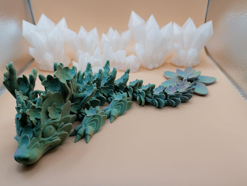May include: A teal and green dragon sculpture with floral details and a gradient finish. The dragon is next to a cluster of white crystal formations and a succulent-like plant. The objects are on a light tan surface.