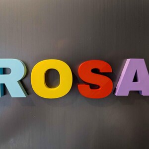 May include: The name "ROSA" spelled out in colorful, three-dimensional letters. The letters are light blue, yellow, red-orange, and purple. The letters are displayed against a gray background.