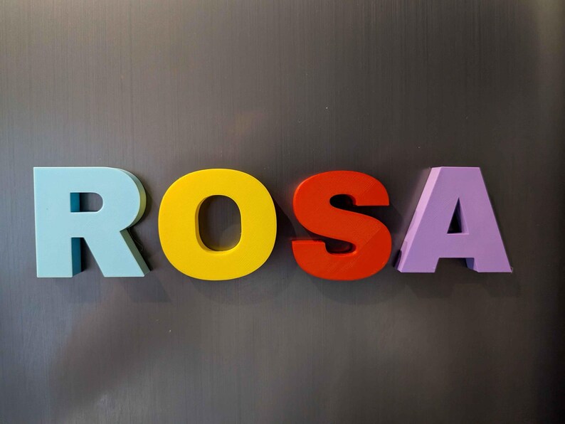 May include: The name "ROSA" spelled out in colorful, three-dimensional letters. The letters are light blue, yellow, red-orange, and purple. The letters are displayed against a gray background.