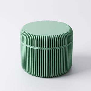 May include: A mint green, cylindrical container with a lid. The container's body features vertical, ridged detailing, and the lid has a flat, textured surface. The design is minimalist and modern, suitable for storage or decorative purposes.