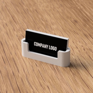 May include: A white business card holder with a black card that reads "COMPANY LOGO" in white text. The holder is made of a light-colored material and sits on a wooden surface.