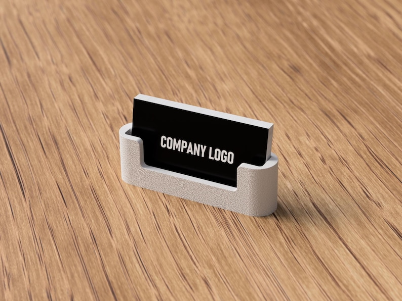 May include: A white business card holder with a black card that reads "COMPANY LOGO" in white text. The holder is made of a light-colored material and sits on a wooden surface.