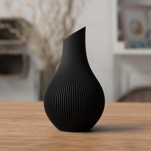 May include: A black, modern vase with a unique, ridged texture. The vase has a curved, organic shape and a slanted top. It is displayed on a wooden table, showcasing its elegant design. The vase is approximately 10 inches tall.