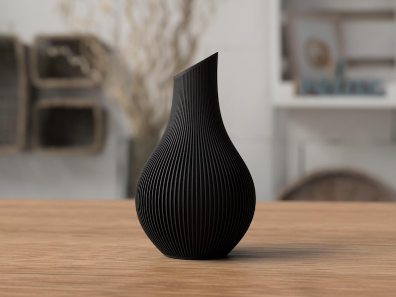 May include: A black, modern vase with a unique, ridged texture. The vase has a curved, organic shape and a slanted top. It is displayed on a wooden table, showcasing its elegant design. The vase is approximately 10 inches tall.