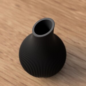 May include: A black, modern vase with a rounded base and a flared, angled opening. The vase has a textured, ribbed design and sits on a wooden surface. The vase is likely used for decorative purposes.