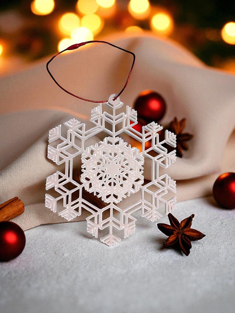 May include: A white snowflake ornament with intricate details hangs from a red string. The ornament is surrounded by red Christmas ornaments, star anise, and a cinnamon stick. The background features soft, blurred lights.
