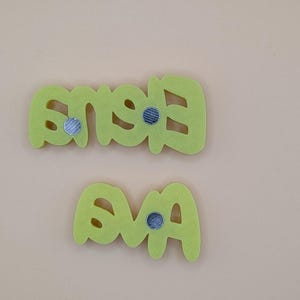 May include: Two lime green felt name magnets spelling "Eva" and "Eves" with silver magnets on the back. The letters are in a playful, rounded font. The background is a solid, light beige colour.