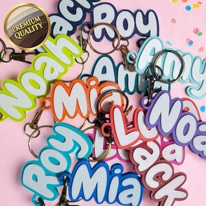 May include: A collection of colorful name keychains with white outlines and metal key rings. The names include "Noah", "Mia", "Roy", "Amy", and "Jack". The keychains are in various colors, including green, orange, blue, and purple.