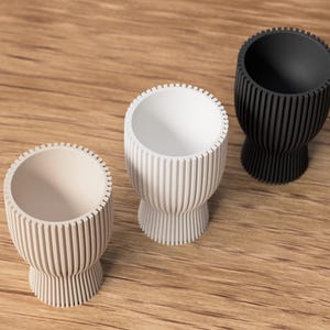 Pu&ograve; includere: Tre vasi decorativi moderni in colori neutri. I vasi sono cilindrici con dettagli a coste verticali e una base svasata. Uno &egrave; bianco sporco, uno bianco e il terzo nero. Sono su una superficie di legno.