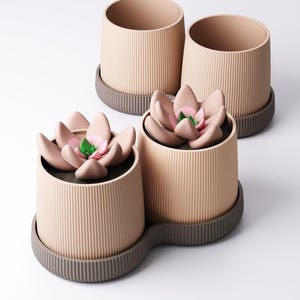 May include: Three beige ceramic plant pots with vertical ribbed detailing. Two are empty, and one contains a faux succulent with pink petals and a green centre. The pots sit on a dark grey, ridged base.
