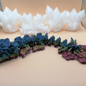 May include: An assortment of decorative crystal formations. The image features several white, spiky crystal clusters and a segmented, multi-colored sculpture with blue, green, and purple hues. The objects are arranged on a light-colored surface.
