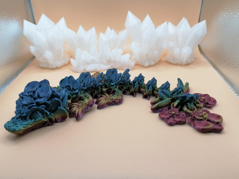 May include: An assortment of decorative crystal formations. The image features several white, spiky crystal clusters and a segmented, multi-colored sculpture with blue, green, and purple hues. The objects are arranged on a light-colored surface.