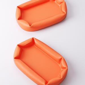 May include: Two orange, rectangular trays with rounded corners are displayed against a white background. The trays have a soft, puffy appearance, with the edges slightly gathered. The color is a vibrant, solid orange.