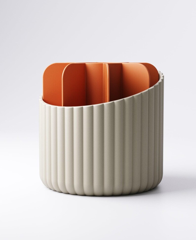 May include: A cylindrical desk organizer with a ribbed, off-white exterior and an orange interior with vertical dividers. The organizer has a curved top edge and a minimalist design, suitable for office or home use.
