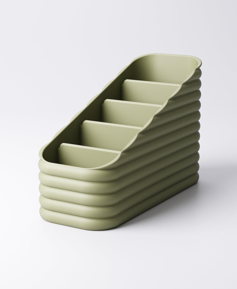 May include: A sage green desk organizer with a stepped design. The organizer has four compartments and a ribbed exterior. The overall shape is rectangular with rounded edges. The product is made of a solid, matte material.