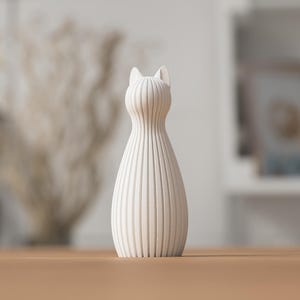 Puede incluir: Un jarr&oacute;n decorativo con forma de gato, de color crema. El jarr&oacute;n tiene un cuerpo delgado y acanalado y orejas puntiagudas. Se encuentra sobre una superficie de madera. El fondo est&aacute; borroso, con indicios de otra decoraci&oacute;n.