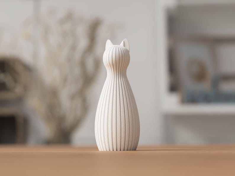 Puede incluir: Un jarr&oacute;n decorativo con forma de gato, de color crema. El jarr&oacute;n tiene un cuerpo delgado y acanalado y orejas puntiagudas. Se encuentra sobre una superficie de madera. El fondo est&aacute; borroso, con indicios de otra decoraci&oacute;n.