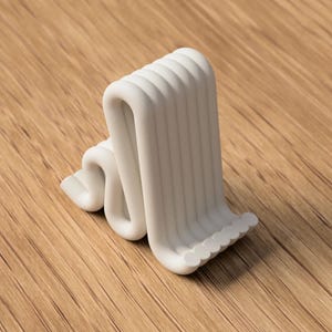 Puede incluir: Una escultura abstracta blanca con un dise&ntilde;o ondulado en forma de cinta. La escultura tiene una serie de crestas verticales y redondeadas en un lado y una serie de peque&ntilde;as formas redondeadas en la base. El objeto est&aacute; sobre un fondo de madera.