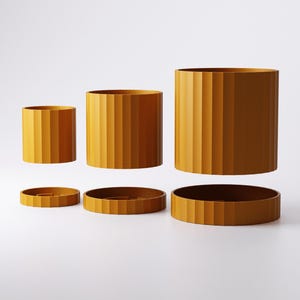 Puede incluir: Tres macetas cil&iacute;ndricas de color amarillo mostaza con texturas acanaladas verticales, cada una acompa&ntilde;ada de una base circular a juego. Las macetas est&aacute;n dispuestas en fila, aumentando de tama&ntilde;o de izquierda a derecha, con cada maceta suspendida sobre su base.