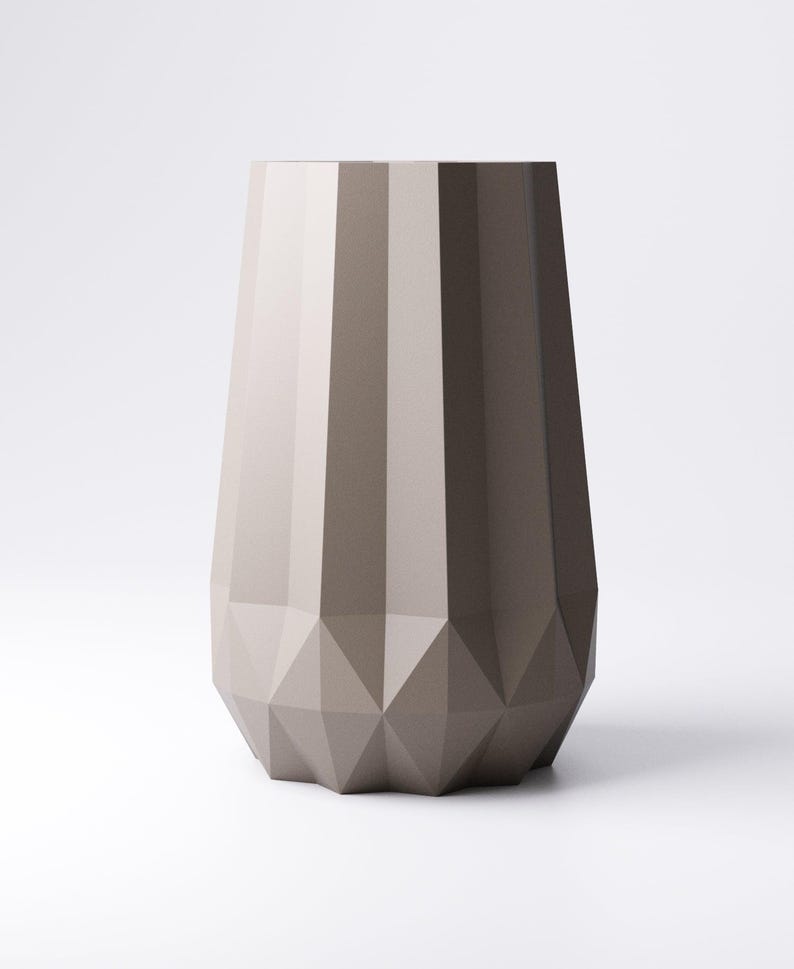 K&ouml;nnte beinhalten: Eine hohe, geometrische Vase in Graut&ouml;nen. Die Vase hat ein facettiertes Design am Boden und vertikale Rippen am K&ouml;rper. Die neutrale Farbpalette und die moderne Form machen sie zu einem vielseitigen Dekorationsst&uuml;ck.