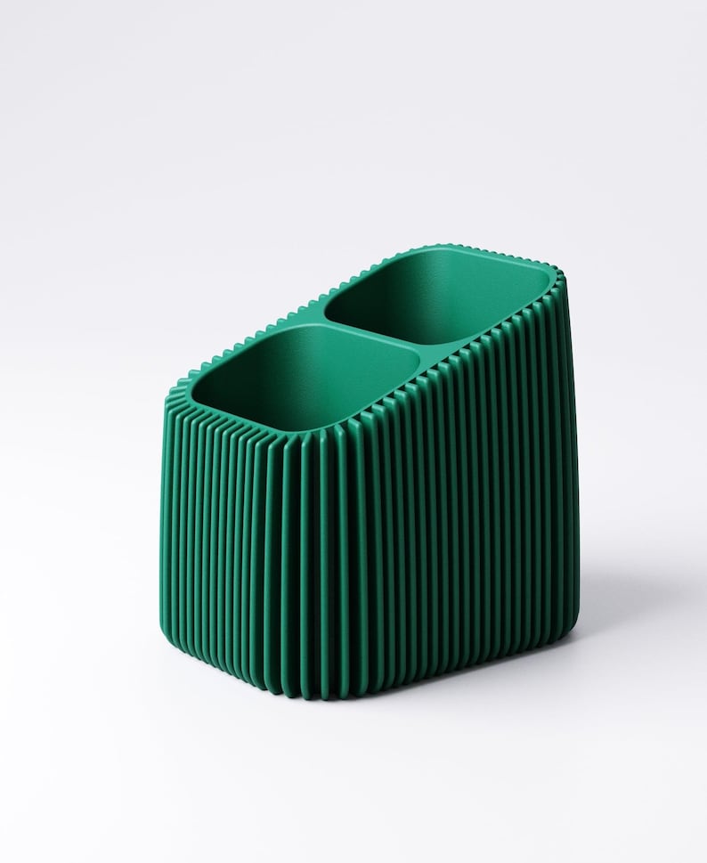 May include: A green desk organizer with two compartments. The organizer has a ribbed exterior and a rectangular shape. The interior is smooth, and the exterior has vertical lines. The organizer is made of a solid, opaque material.
