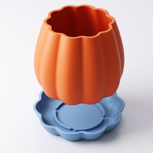 May include: An orange pumpkin-shaped container with a scalloped rim sits atop a light blue, flower-shaped base. The container has vertical ridges and a smooth, matte finish. The base has a circular indentation in the center.